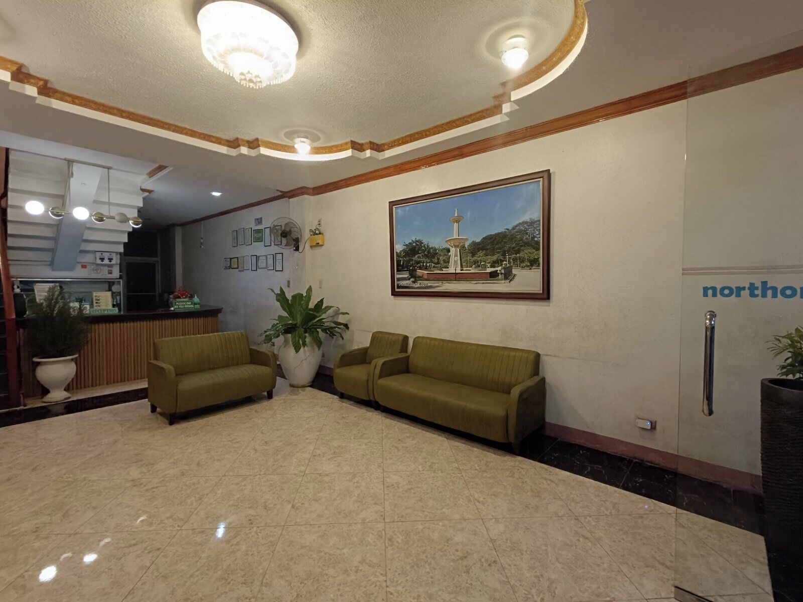 A lobby with a reception desk, two sofas, a large potted plant, and a framed picture of a fountain.