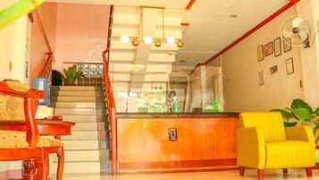 A hotel lobby with a reception desk, a staircase, a yellow armchair, and a fan on the wall.