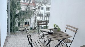 A balcony with a wooden table and chairs, a potted plant, and a view of a building with a tiled roof. - Apartment Bremen Neustadt (Bremen)
