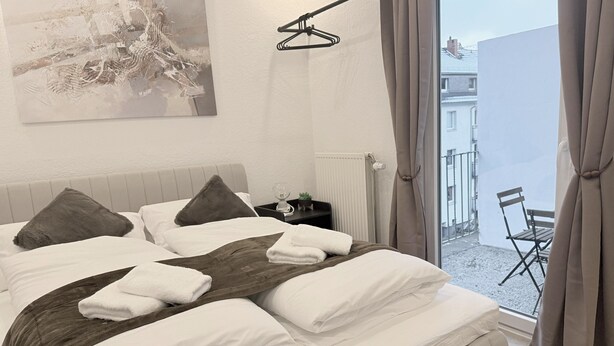 A neatly made bed with white linens and a brown throw blanket, a bedside table with a glass, a small plant, and a hanging light fixture. - Apartment Bremen Neustadt (Bremen)