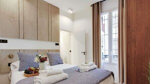 A hotel room with a large bed, a bedside table with a fruit basket and towels, a window with curtains, and a wooden headboard. - Superb apartment with AC - Center of Paris (Paris)