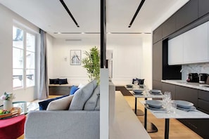 Uma sala moderna com um sofá cinzento, uma mesa de jantar posta para quatro pessoas e uma cozinha com armários escuros e bancada de mármore.