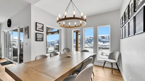 Een ruime eethoek met een houten tafel en stoelen, grote ramen met uitzicht op een besneeuwde berg, en een kroonluchter.