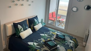 A bedroom with a bed, pillows, a hanging light fixture, and a view of the beach through a sliding door. - PLEINE MER Front de Mer - 1st Et. Elevator - 4P - Parking - Wifi - Linen Ok (Courseulles-sur-Mer)