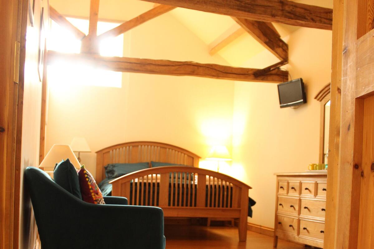 A room with a wooden floor, a bed with a headboard, a wooden dresser, and a television mounted on the wall.