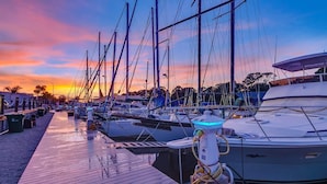 Een jachthaven met aan de kade afgemeerde boten, een houten looppad en een ondergaande zon op de achtergrond.