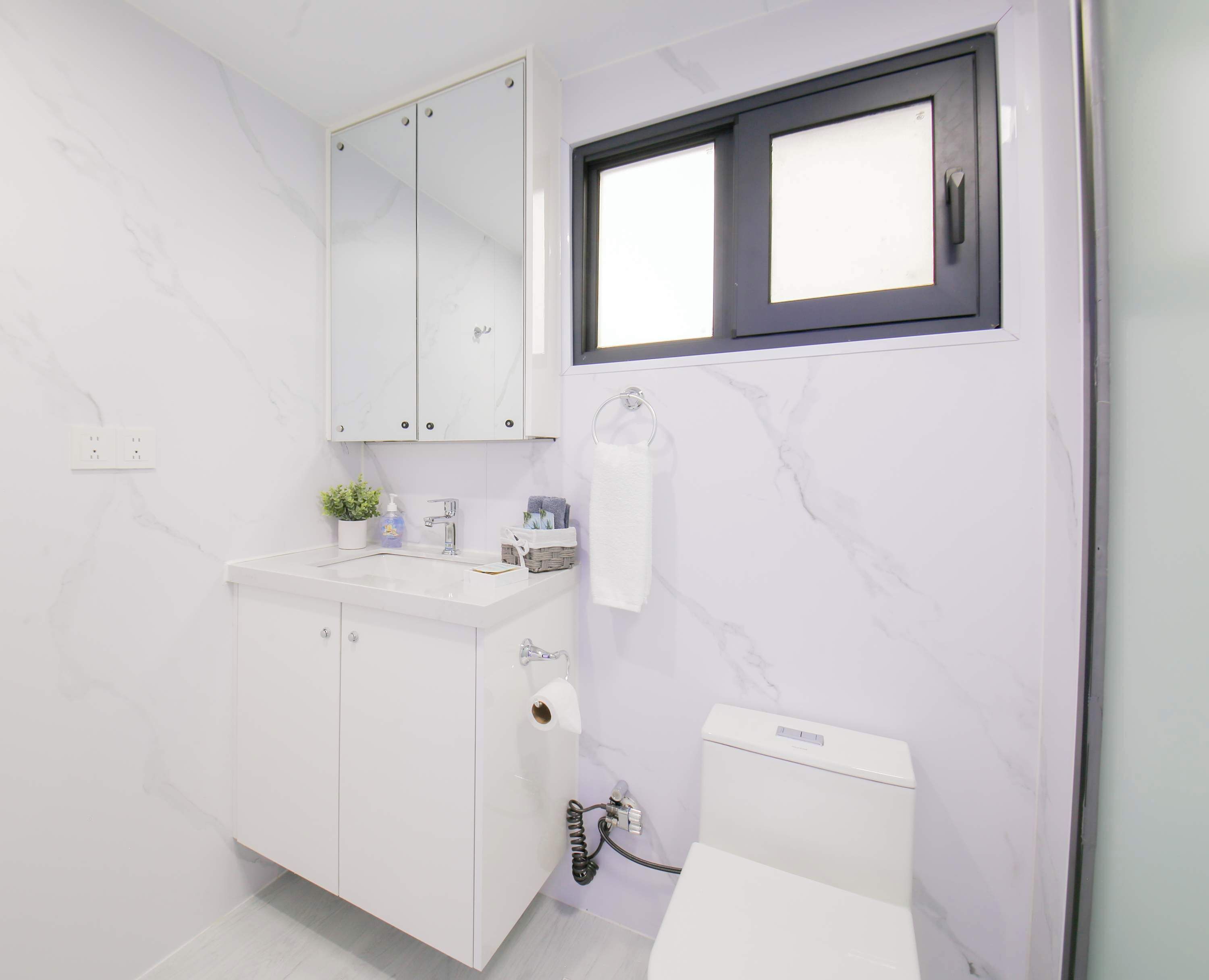 Un baño moderno con pared de mármol blanco, lavamanos blanco, espejo, inodoro y ventana con marco negro.