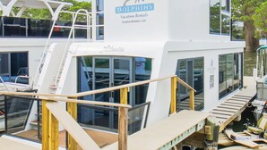 A white boat with 'Dolphins Vacation Rentals' branding, air conditioning units, and a deck with seating. - Avalon 2-Bedroom Floating Home (Merritt Island)