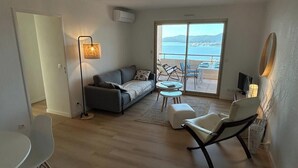 Una sala de estar moderna con un sofá, una mesa de centro y un sillón. Hay un ventanal corredero que da a un balcón con vistas al mar.