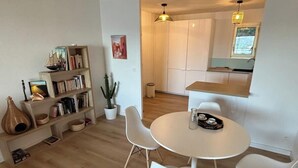 Una sala de estar moderna con una mesa redonda blanca y cuatro sillas blancas, una estantería con libros y objetos decorativos, una planta en maceta y una ventana con vistas al área de la cocina.