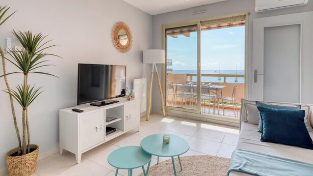 A modern living room with a flat-screen TV, a white cabinet, two small round tables, a sofa with blue cushions, and a large sliding glass door leading to a balcony with a view of the sea. - Exceptional sea view in Lavandou (Le Lavandou)