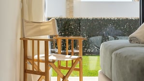 Een kamer met een bed, een houten stoel en een kleine tafel. Er is een raam met gordijnen, een uitzicht op een tuin en een waterpartij buiten.