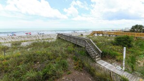 A wooden boardwalk leading to a beach with a clear view of the ocean and a distant pier.
