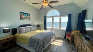 Een slaapkamer met een bed, een plafondventilator, een raam met uitzicht op zee, een dressoir en een nachtkastje met een lamp.