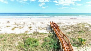 Een strand met een houten boardwalk naar de kust, zandduinen en een helderblauwe lucht.