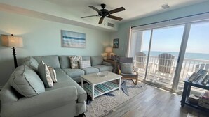 Een woonkamer met een hoekbank, een salontafel en een fauteuil. Er is een plafondventilator, een groot raam met uitzicht op het strand en een balkon met tuinmeubels.