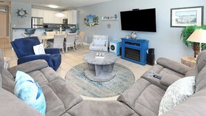 A living room with a grey sofa, a blue armchair, and a coffee table. There is a kitchen in the background with white cabinets and a blue countertop. - 2nd Row 4 BR Beach House with a Shared Pool! - Chubby's Anchor (Surfside Beach)
