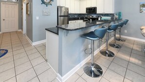 A kitchen with white cabinets, a stainless steel refrigerator, a microwave, and a granite countertop. There are four blue bar stools and a decorative clock on the wall. - 2nd Row 4 BR Beach House with a Shared Pool! - Chubby's Anchor (Surfside Beach)