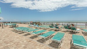 A beachside lounge area with rows of green lounge chairs and tables, a pool, and a view of the ocean.