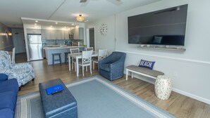 A modern living room with a sofa, ottoman, and a coffee table. There is a dining area with chairs and a table, and a kitchen in the background.