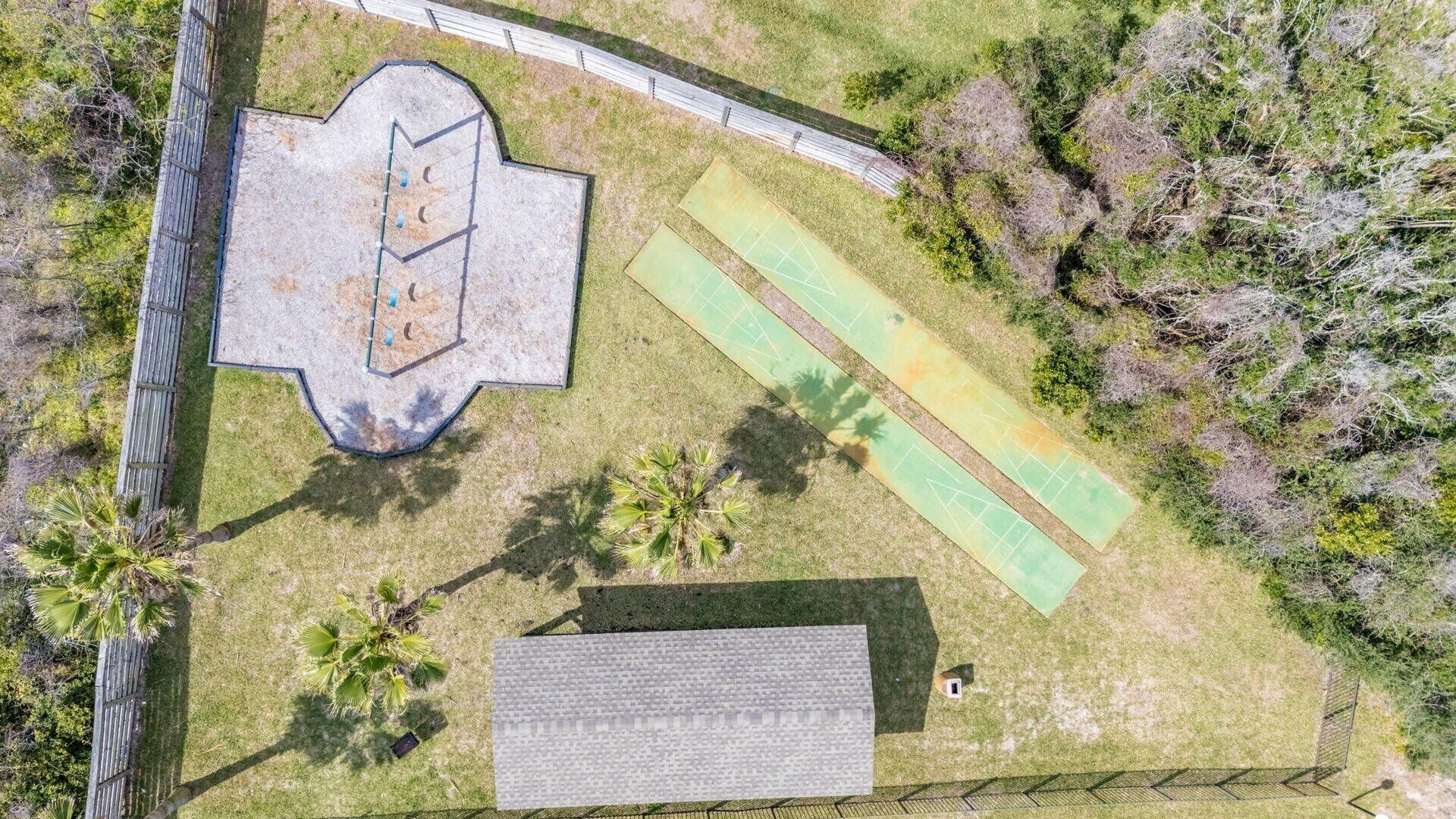 An aerial view of a property with a large, irregularly shaped building, a fenced area with a basketball court, and a small structure with a sloped roof.