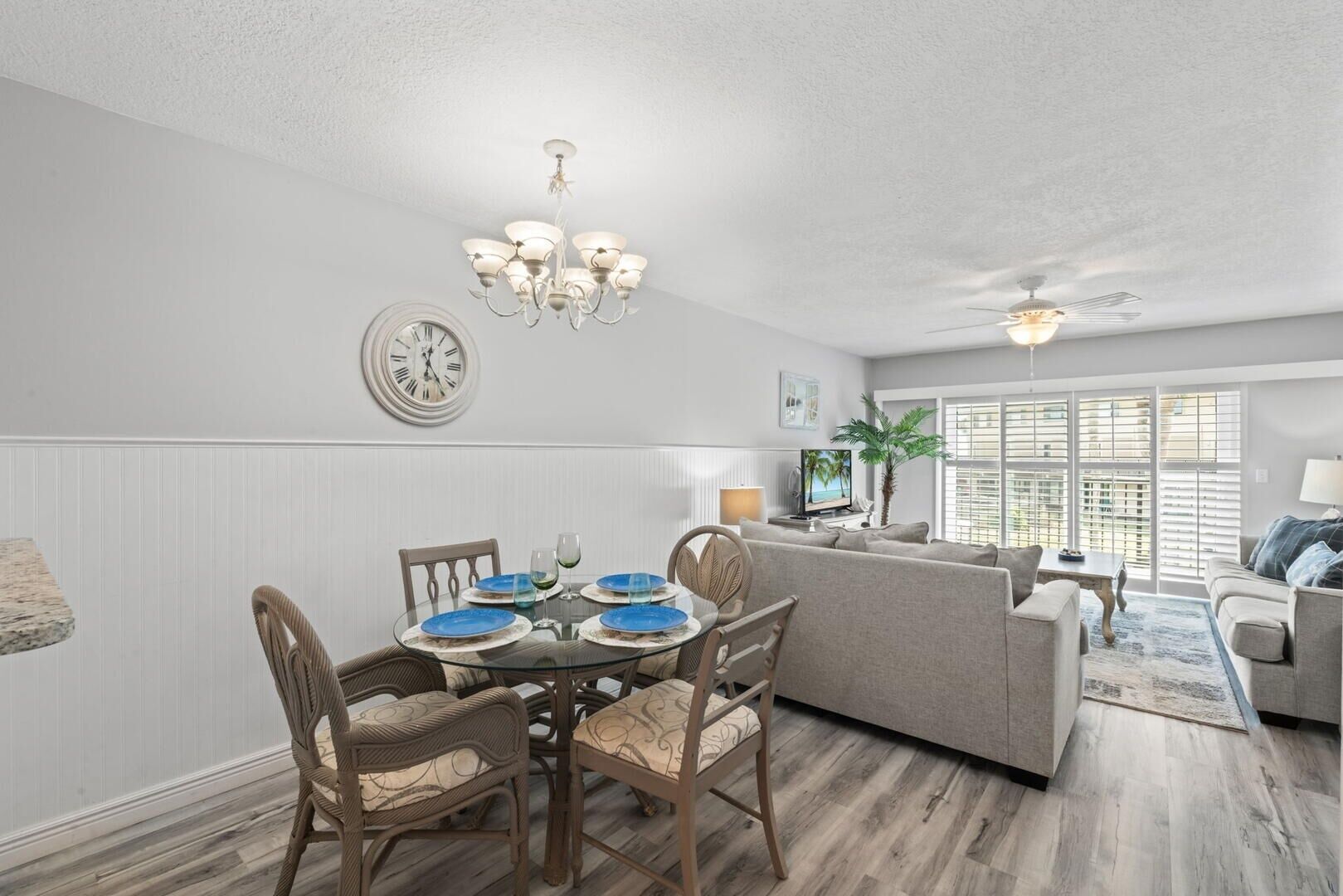 A modern living room with a dining area, featuring a round glass table, wooden chairs, a grey sofa, and a ceiling fan.