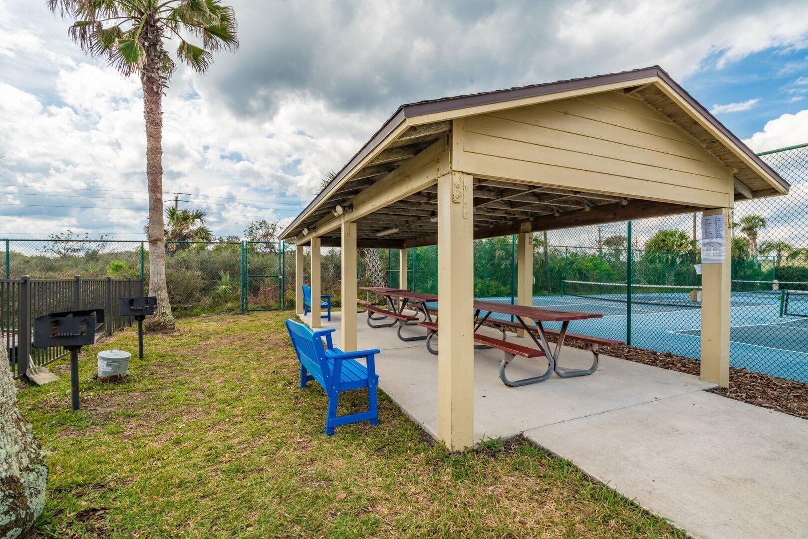 A covered picnic area with benches and picnic tables, surrounded by a fence and palm trees.