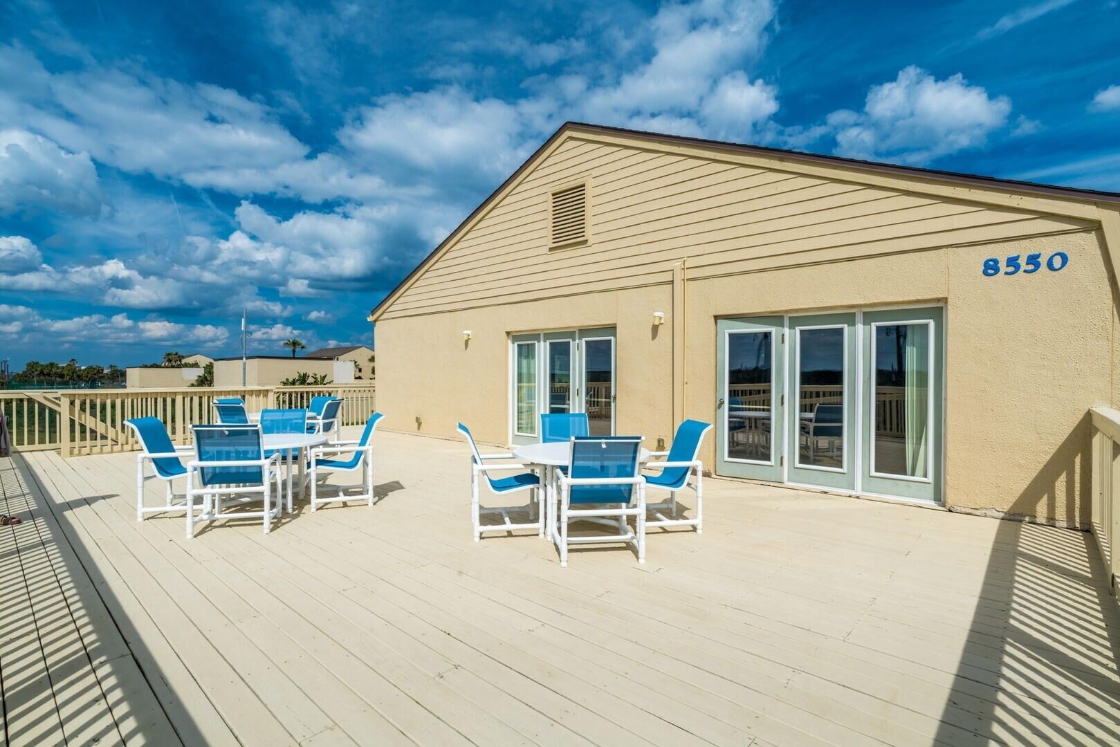 Ein Hotel mit der Nummer 8550 an der Seite, eine überdachte Terrasse mit blauen Stühlen und Tischen sowie ein klarer, blauer Himmel.