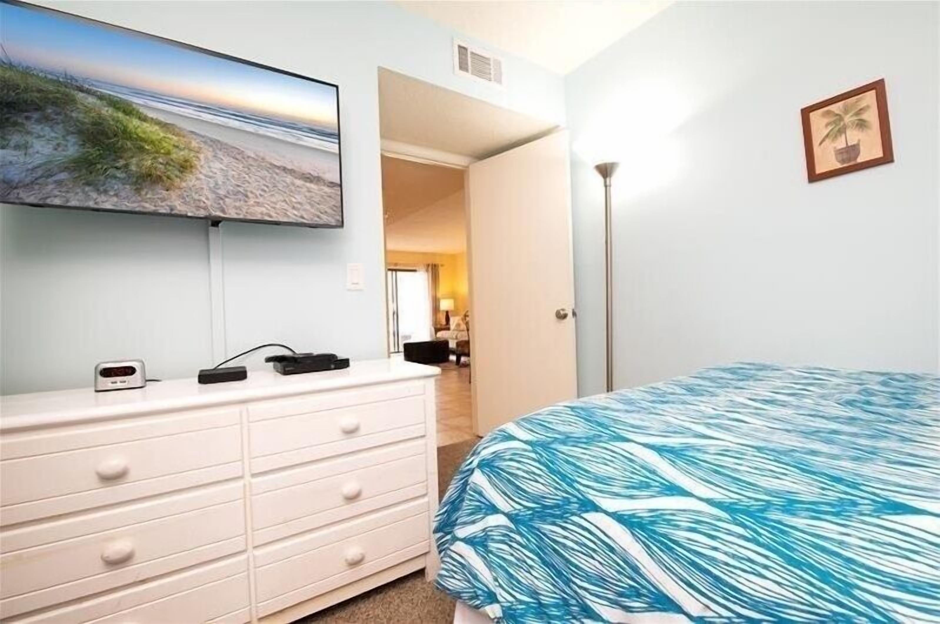 A bedroom with a bed, a dresser, a flat-screen TV, and a door leading to another room.