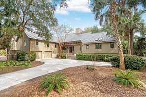 A two-story house with a front porch, a driveway, and a landscaped garden with palm trees and shrubs. - Stunning view in great Vanderhorst area location (Kiawah Island)