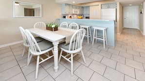 A dining area with a white table and chairs, a kitchen with light blue cabinets, and a white door in the background. - Oceanfront 3 BR Condo with a Shared Pool! Surf Harbor 203 (Surfside Beach)