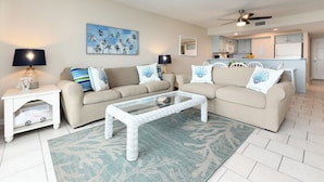 A living room with two beige sofas, a glass coffee table, and a patterned rug. There are two white nightstands with lamps, a ceiling fan, and a kitchen area in the background. - Oceanfront 3 BR Condo with a Shared Pool! Surf Harbor 203 (Surfside Beach)