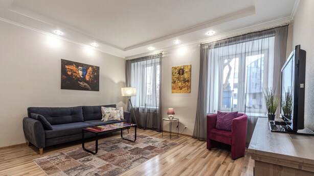 Una sala de estar moderna con un sofá, una mesita y una silla. Hay dos ventanas con cortinas, un televisor de pantalla plana y una lámpara sobre una mesita auxiliar.