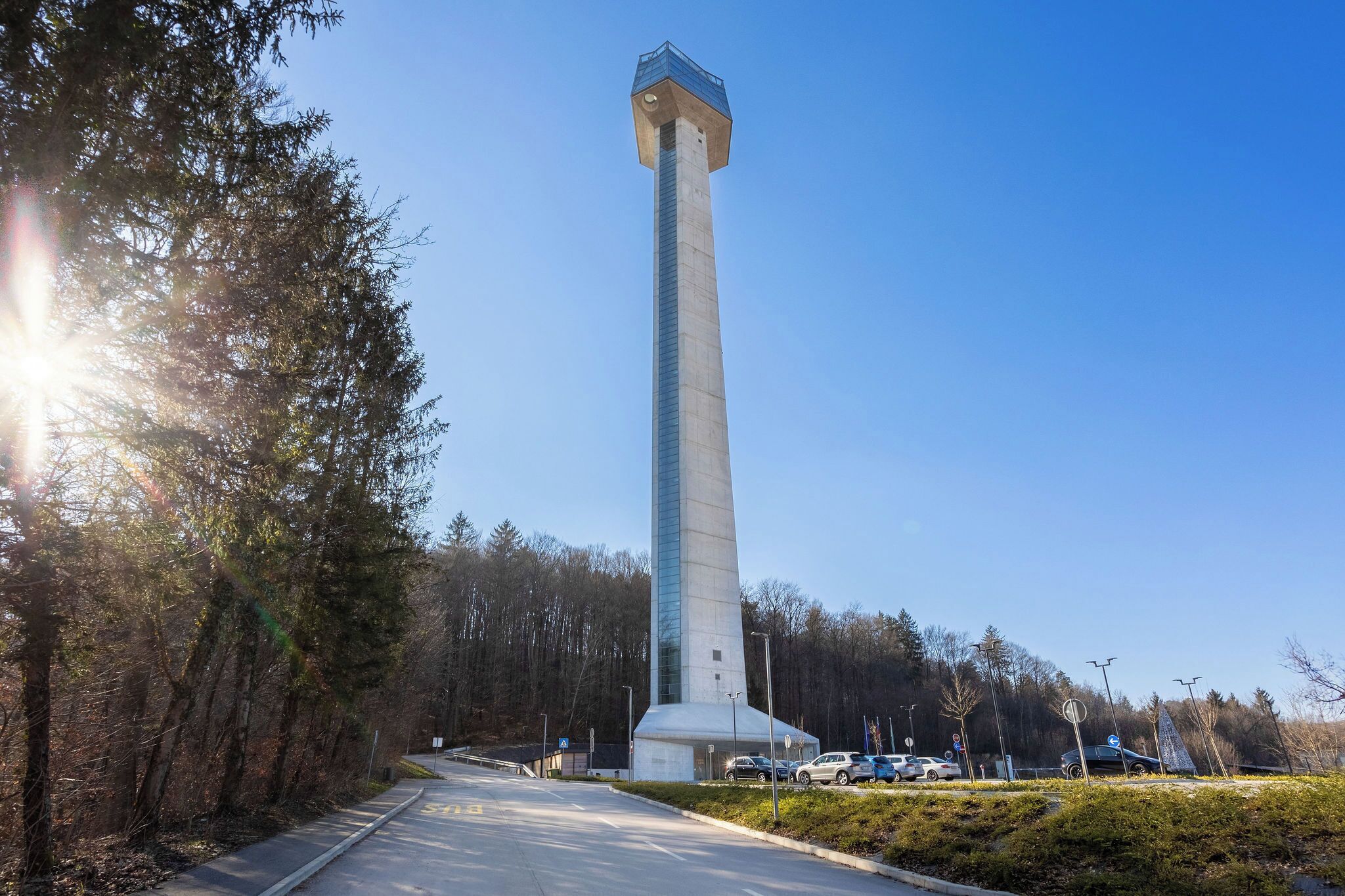 A tall, narrow tower with a flat top, surrounded by trees and a paved road.