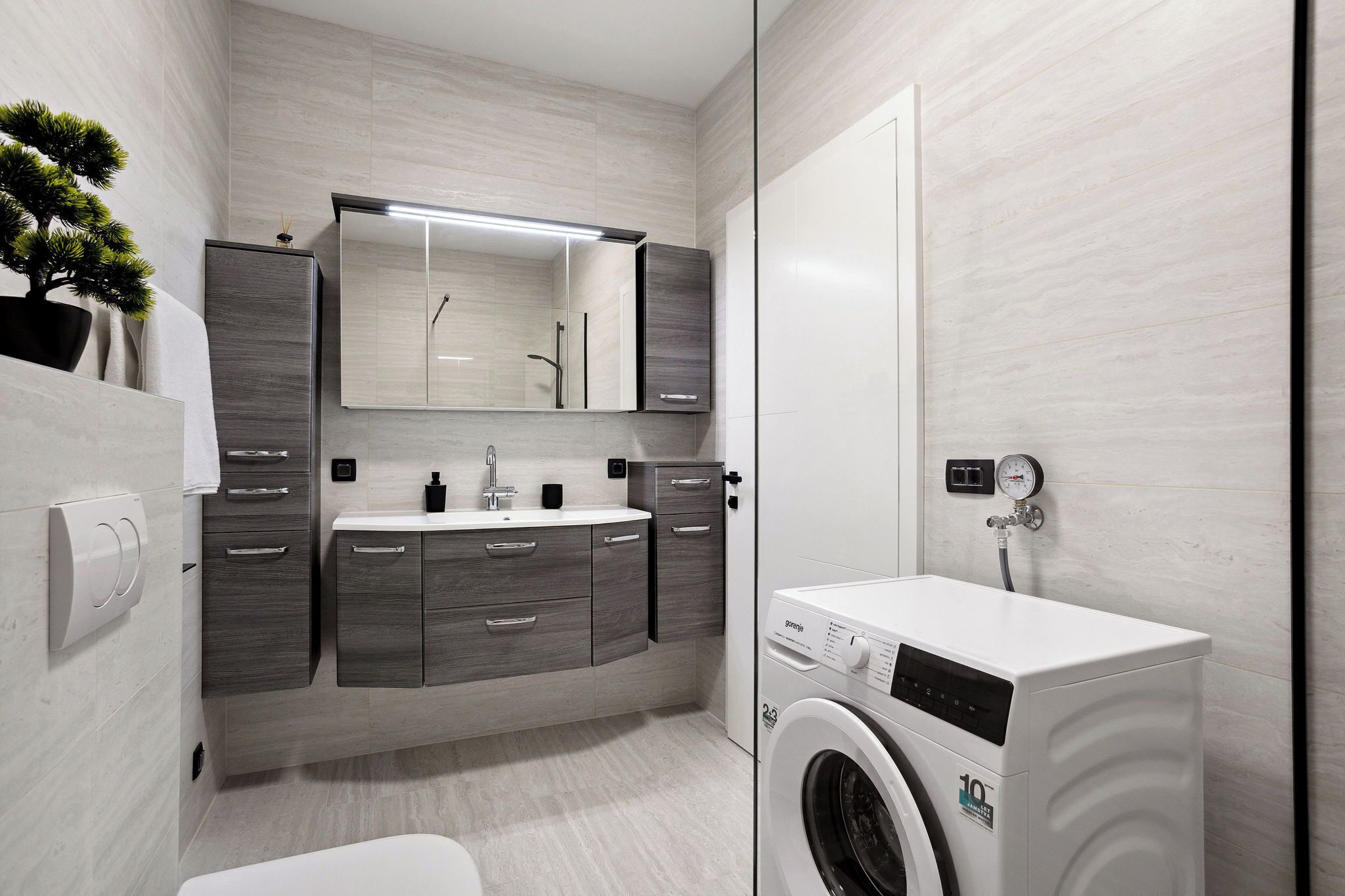 A modern bathroom with grey cabinetry, a built-in sink, a mirror, a washing machine, and a potted plant.