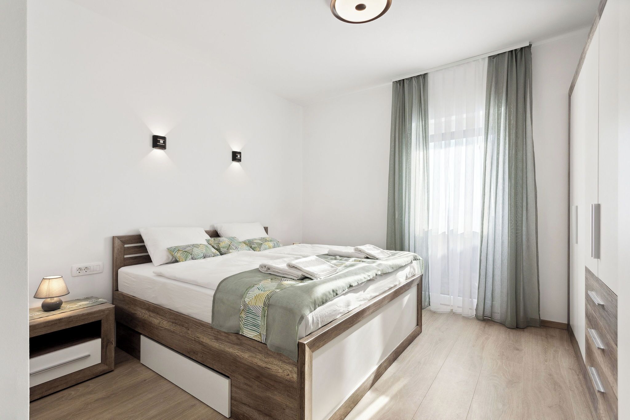 Een moderne slaapkamer met een houten bed, nachtkastjes en een groot raam met gordijnen.
