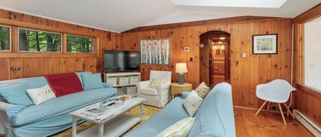 A cozy living room with a sofa, armchair, and a coffee table. There is a television and a lamp on a side table.