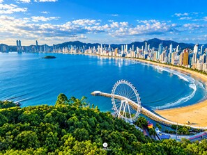 A coastal cityscape with a prominent Ferris wheel, a beach, and a dense urban skyline.