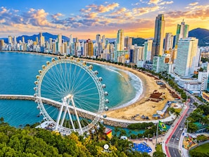 A coastal cityscape with a prominent Ferris wheel, high-rise buildings, and a beach. - Ap Punta Cana em Balneário Camboriú - SC (Balneário Camboriú)