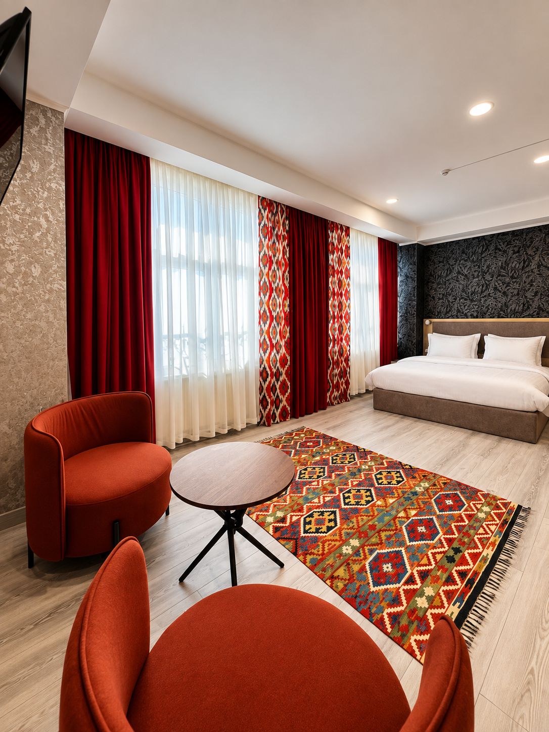 Una habitación de hotel con una cama, dos sillones rojos, una mesita redonda y una alfombra estampada.