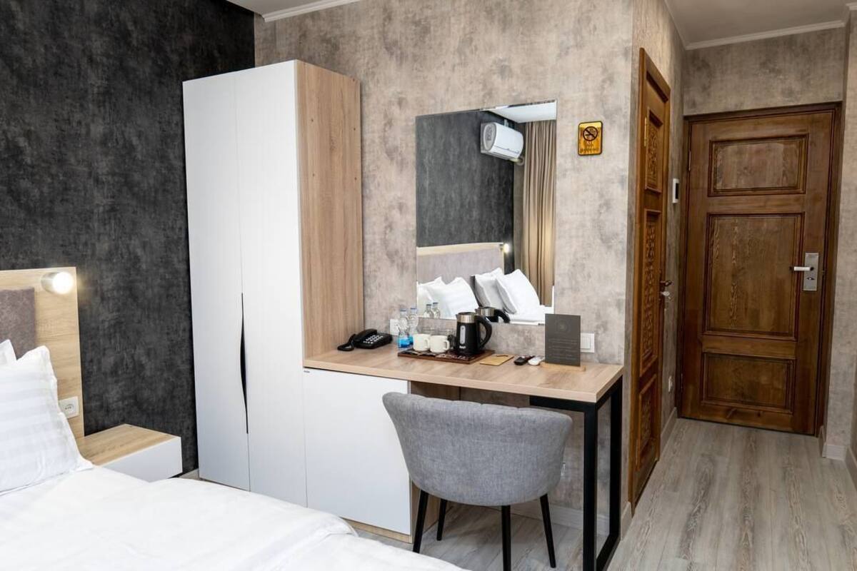 A hotel room with a bed, a desk with a chair, a wardrobe, a mirror, a door, and a small table with a kettle and cups.