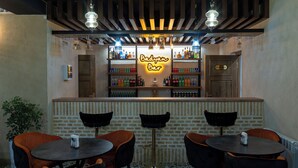 A bar area with a neon sign reading "Badragan Bar", shelves stocked with various bottles, and three round tables with chairs. - Badyan Hotel (Bukhara)