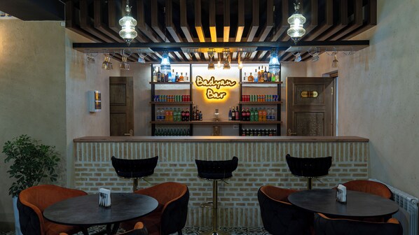 Un espace bar agrémenté d’un panneau néon indiquant « Badragan Bar », d’étagères remplies de différentes bouteilles et de trois tables rondes avec des chaises.