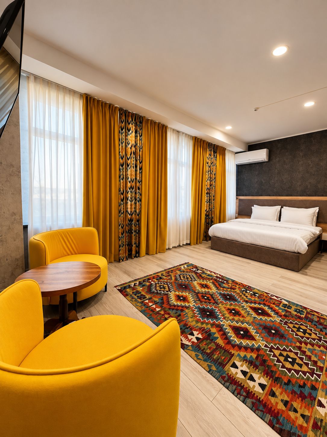 Ein modernes Hotelzimmer mit einem Bett, einem runden Holztisch, zwei gelben Sesseln, einem gemusterten Teppich und einem großen Fenster mit Vorhängen.