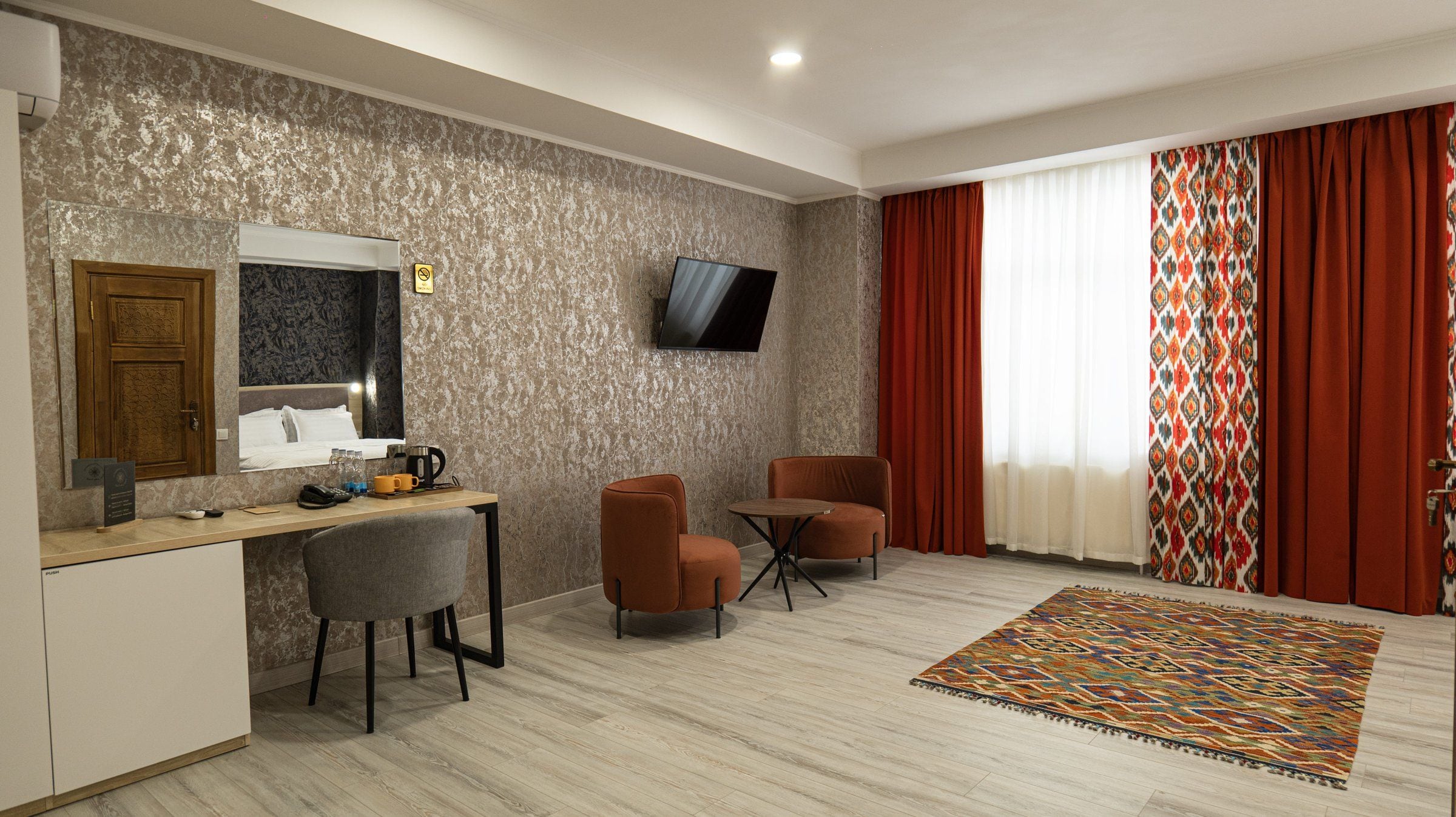 A hotel room with a wooden desk, two chairs, a TV mounted on the wall, a patterned rug, and a large window with red curtains.