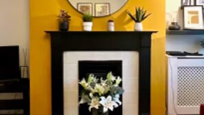 A living room with a fireplace, a yellow accent wall, and a round mirror above it. - Woodcrest Retreat Sleeps 4 With Garden (Reigate)