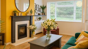 A living room with a fireplace, a large mirror, a green sofa, and a window with a view of greenery. - Woodcrest Retreat Sleeps 4 With Garden (Reigate)
