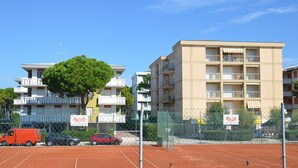 A multi-story residential building with balconies, a fenced tennis court, and parked cars. - Stylish retreat steps from everything (Bibione)