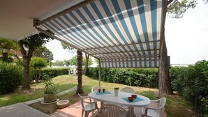 Eine Terrasse im Freien mit einem gestreiften Sonnenschirm, einem runden Tisch, weißen Kunststoffstühlen und einem kleinen Blumentopf.