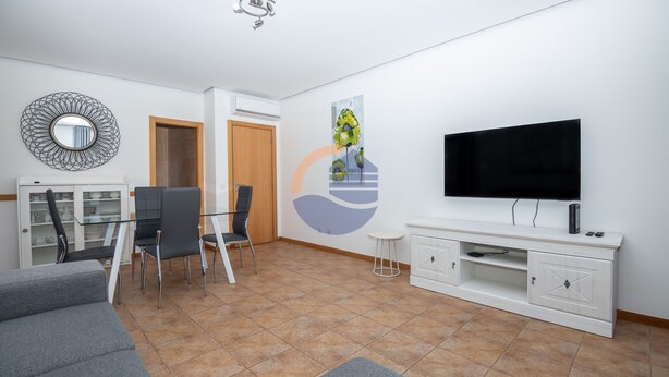 Egy modern nappali, melyben egy síkképernyős televízió, egy üveglapos étkezőasztal és egy szürke színű kanapé található.